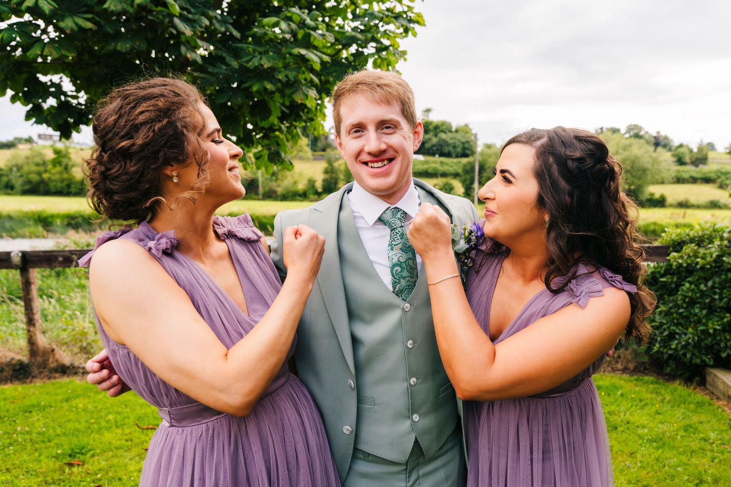 Millhouse slane wedding photographer dublin Bridesmaids in purple dresses playfully adjust groom's tie in garden setting.