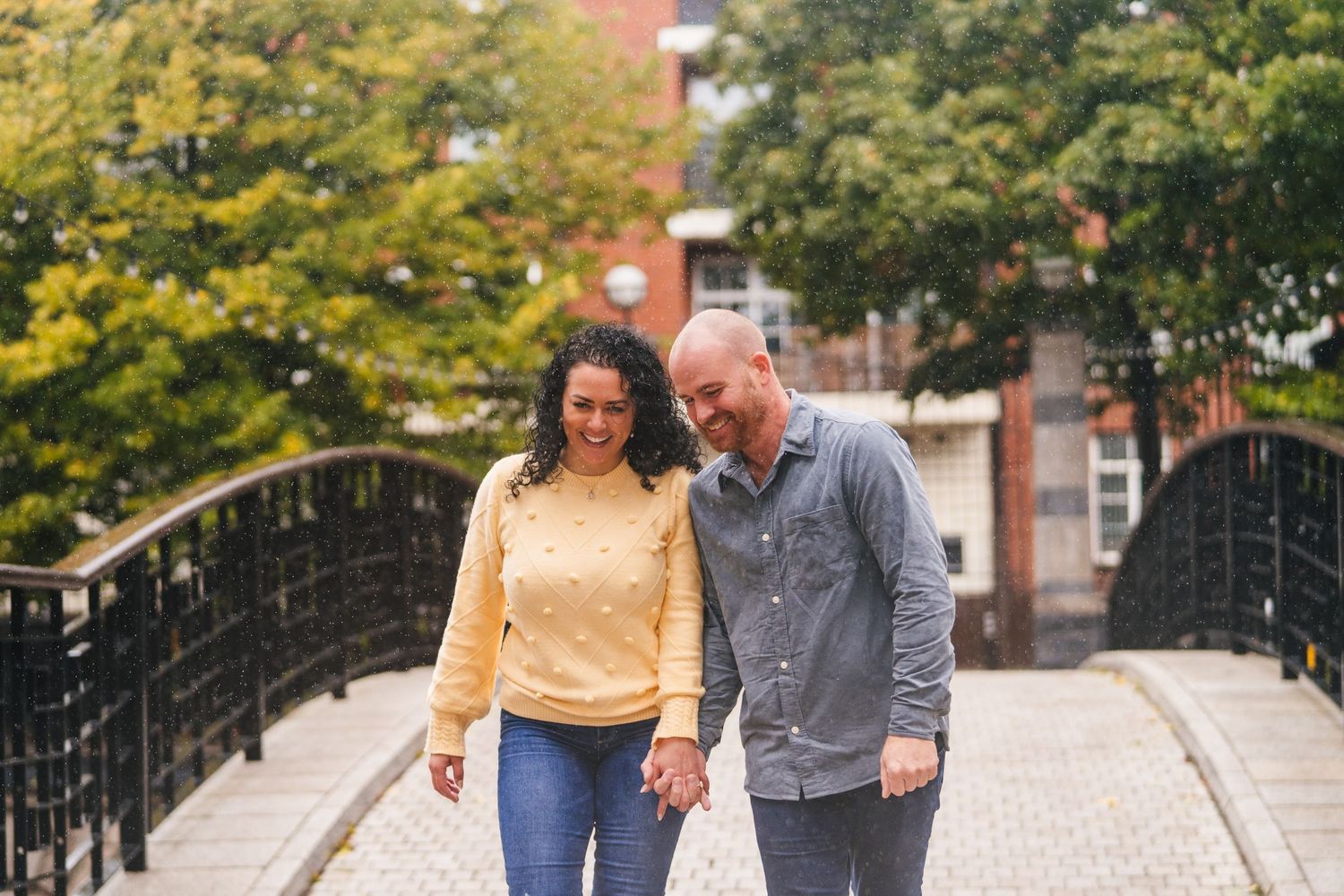A couple walks hand in hand across a brick bridge with autumn trees and red buildings in the background. Proposal Dublin Photographer