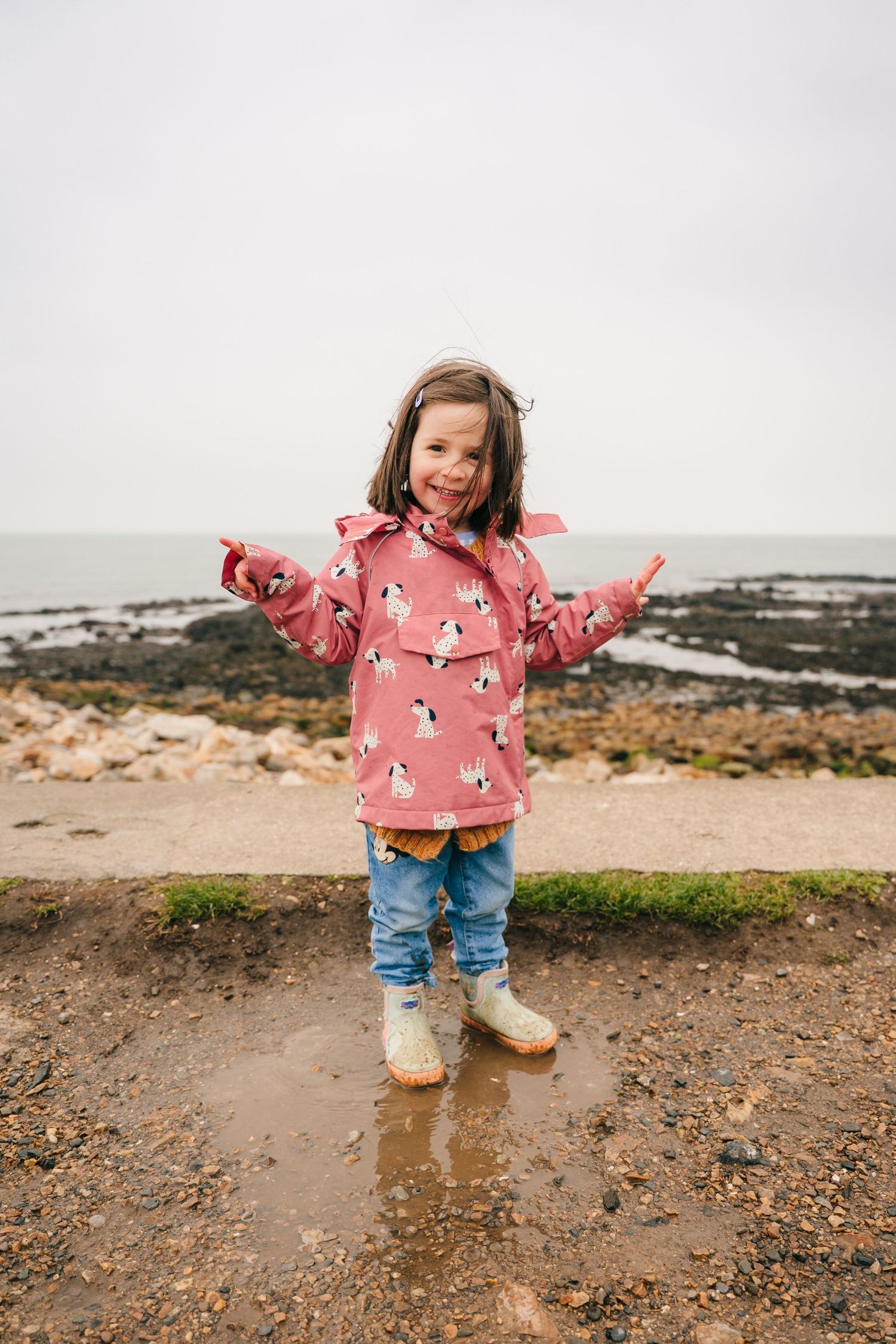 A happy child wearing a pink penguin-patterned raincoat plays in the mud on a cloudy beach day. PHOTOGRAPHER Dublin