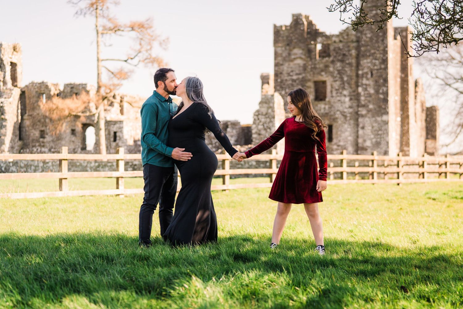 A couple and friend dance together on green field with historic castle ruins in the background on sunny day. Family PHOTOGRAPHER Dublin