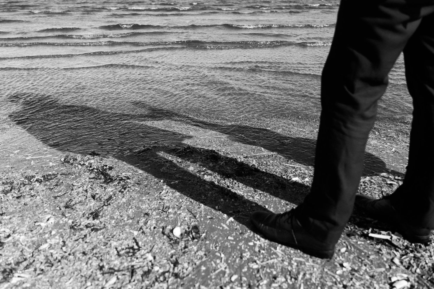 Black and white photo of a long shadow cast on rocky ground near water's edge. PHOTOGRAPHER Dublin