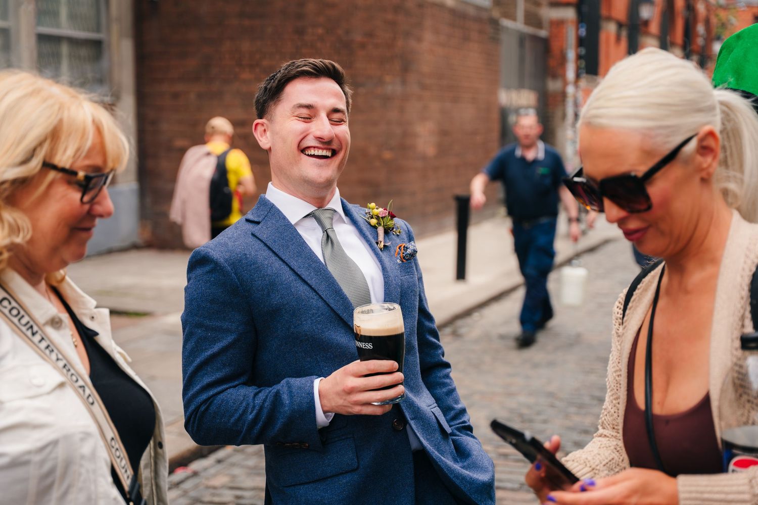 People socializing on a brick-lined city street with someone in a navy suit holding a drink. Same sex wedding PHOTOGRAPHER Dublin