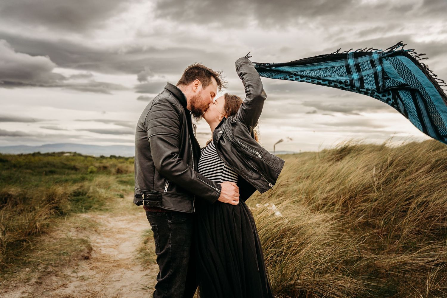A romantic couple share an intimate moment on a windy hillside path under stormy skies. engagement PHOTOGRAPHER Dublin