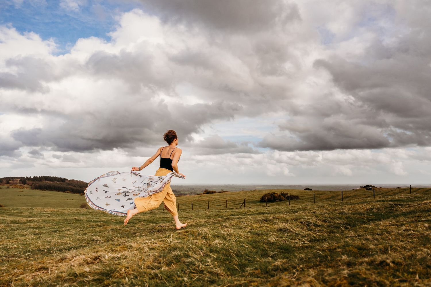 A flowy white dress billows dramatically against a moody sky over rolling green hills and grasslands. Portrait PHOTOGRAPHER Dublin
