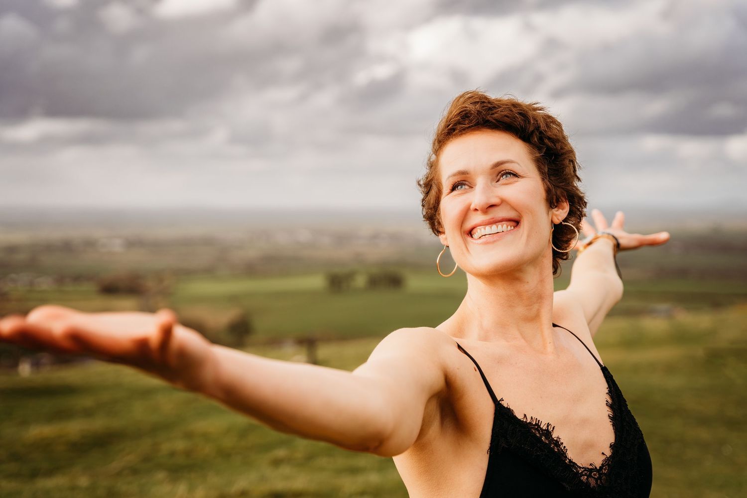 Person in black tank top stretches arms wide while standing on a hilltop overlooking green rolling countryside. Portrait PHOTOGRAPHER Dublin