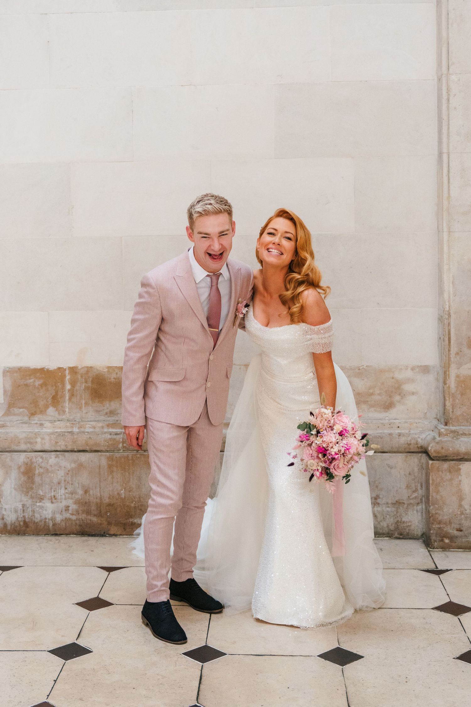Wedding couple in white and pink attire pose playfully against light stone wall with pink bouquet.  city hall dublin wedding photos