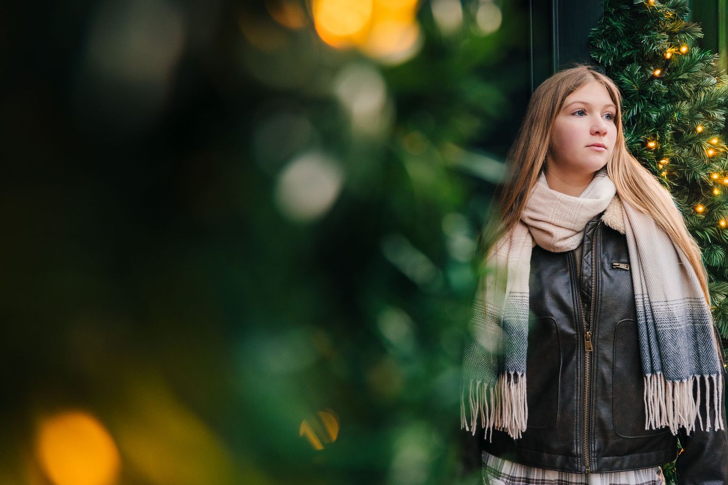 Person in winter scarf and leather jacket poses against blurred holiday lights and greenery background. family PHOTOGRAPHER Dublin