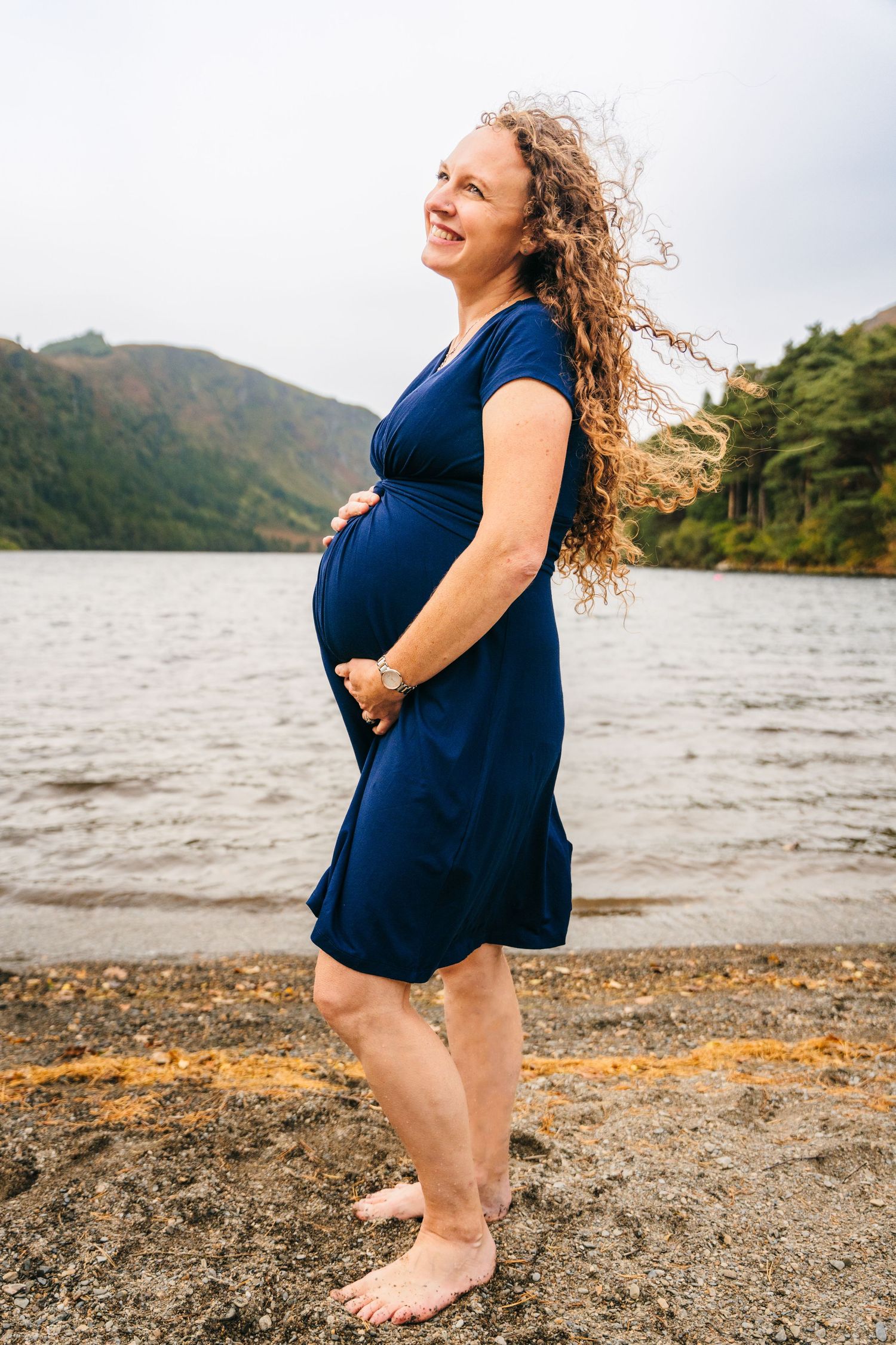 A pregnant woman in a navy dress stands joyfully on a rocky beach with mountains and water in the background.Family photogrAPHER DUBLIN
