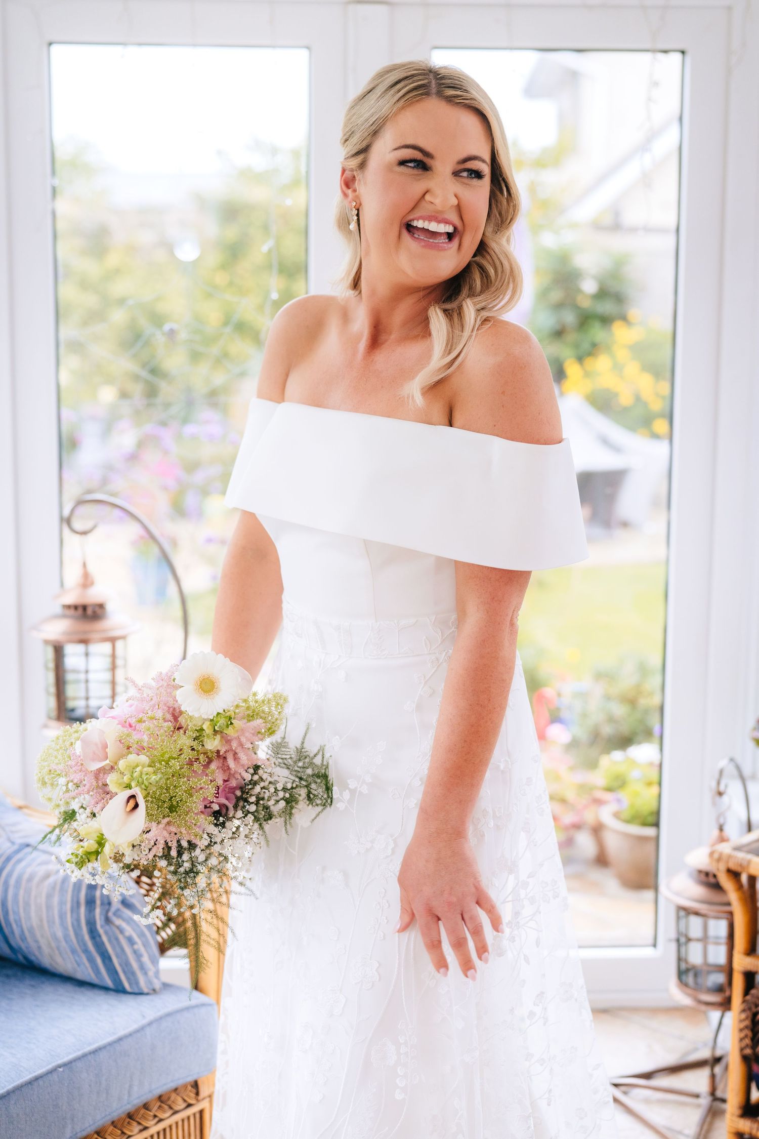 Beautiful bride in an elegant off-shoulder white wedding dress poses joyfully in a bright sunlit room with flowers. The Dean Dublin Wedding