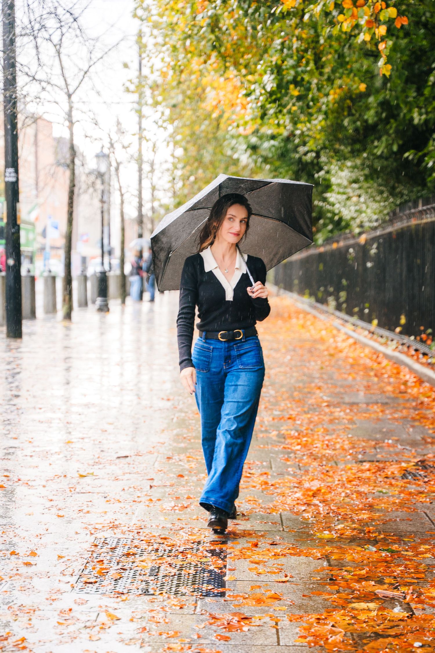 Person with black umbrella walking on autumn sidewalk covered in vibrant orange fallen leaves on rainy day. Headshots Dublin Photographer Po
