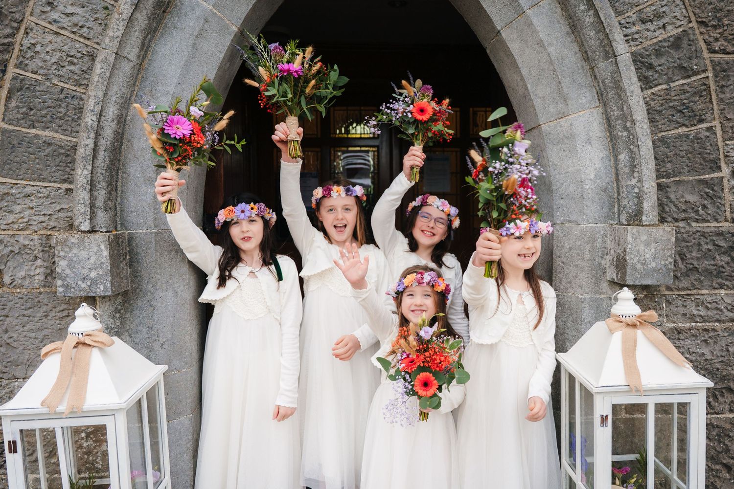 Wedding party in white dresses poses joyfully under a stone archway with pink floral decorations. Wedding PHOTOGRAPHER Dublin ireland