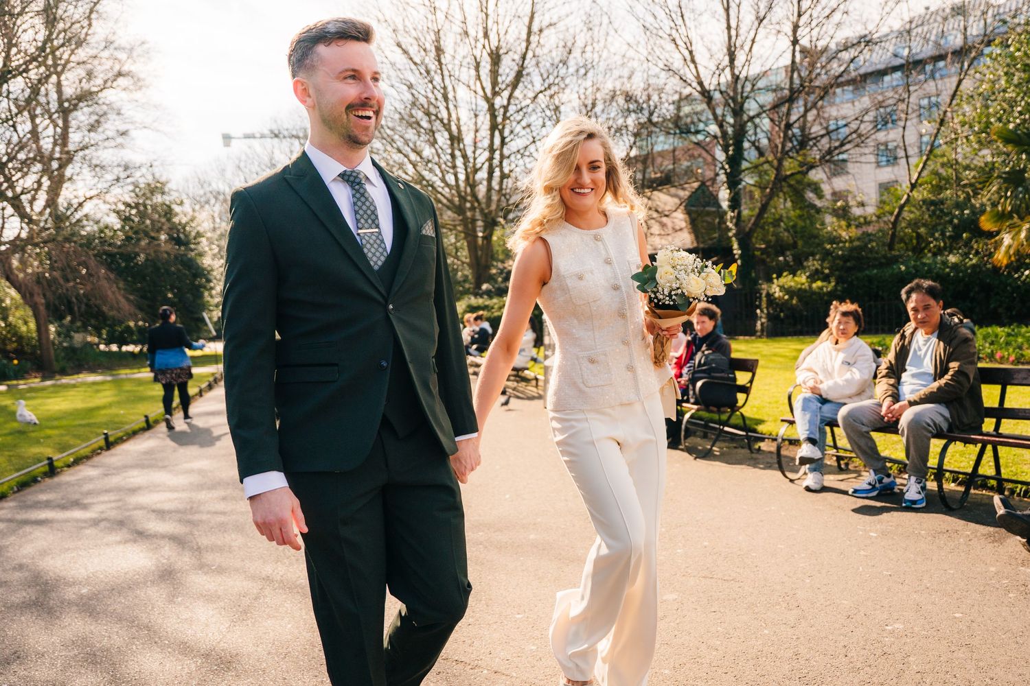 A couple strolls through a sunny park on a spring day, dressed formally in white and dark attire. City Hall Dublin wedding photogrAPher