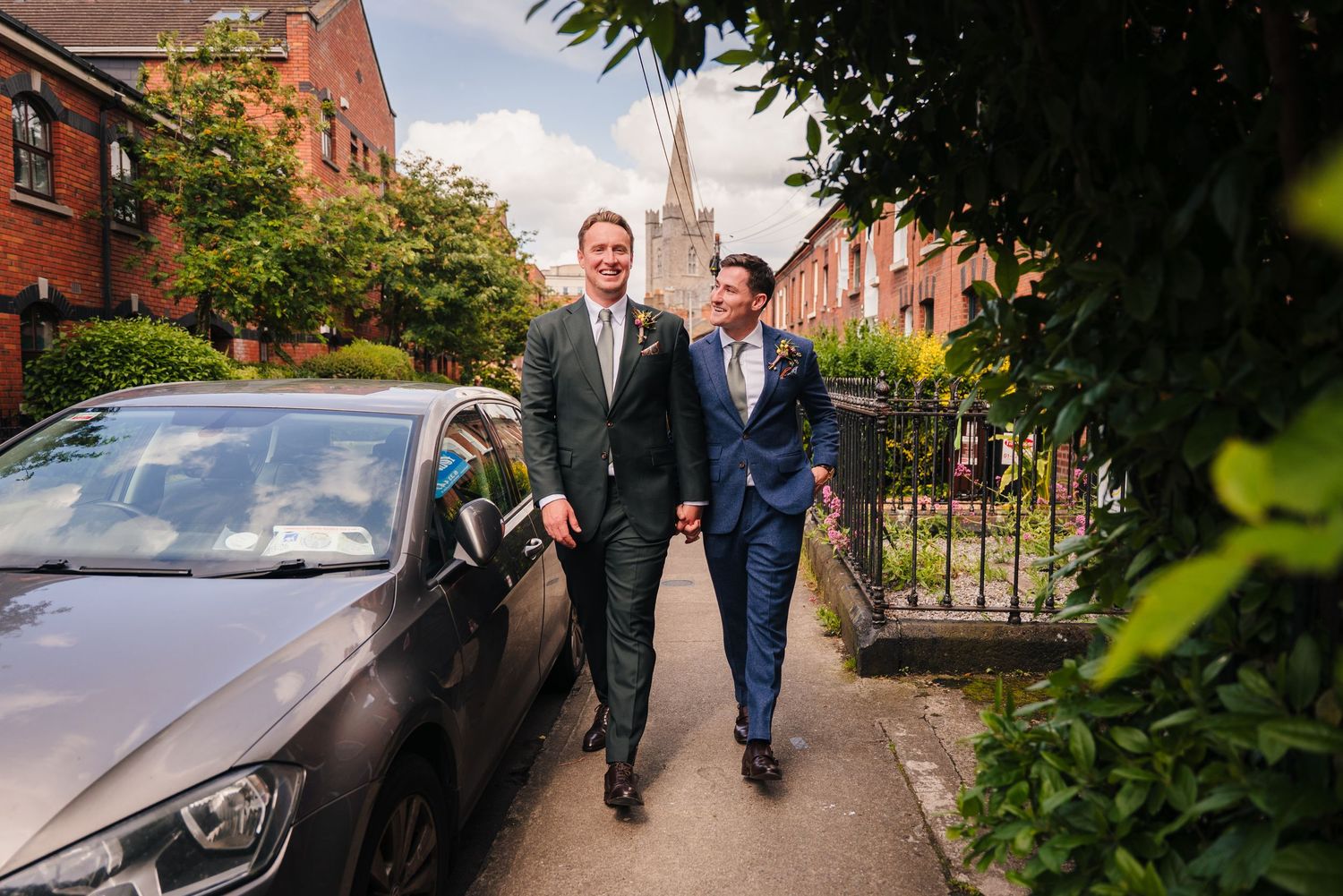 Two people in suits walking together down a residential street with parked cars. Same sex wedding PHOTOGRAPHER Dublin