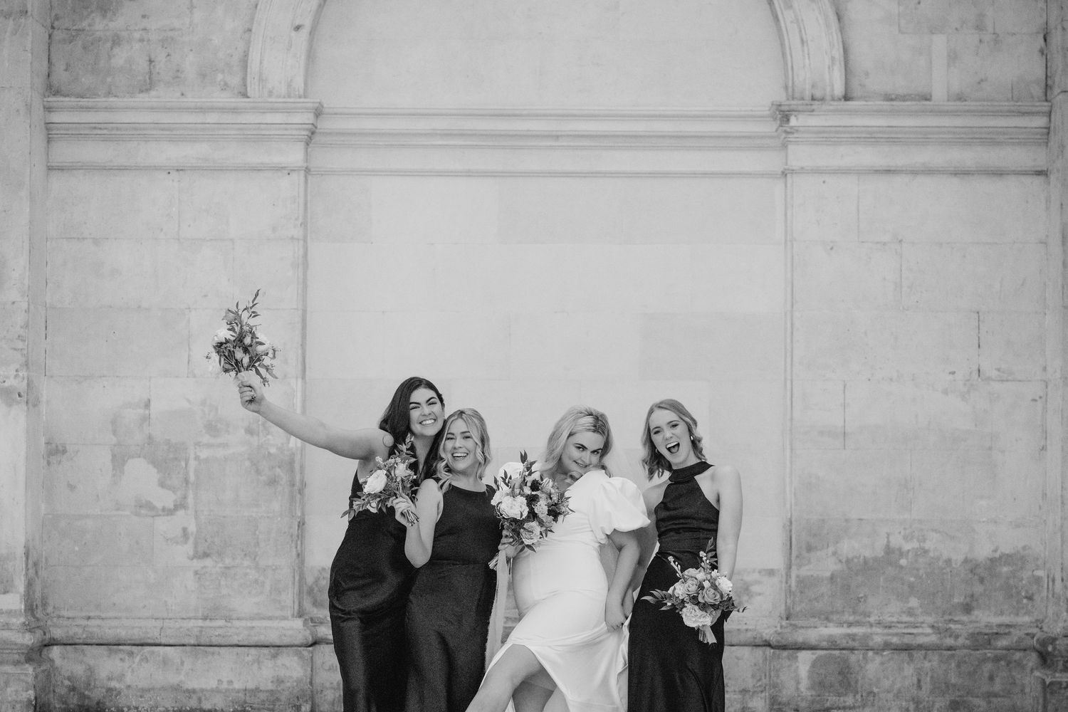 Wedding party jumps in celebration against white stone wall during outdoor photo session at historic venue. City Hall Dublin Photographer