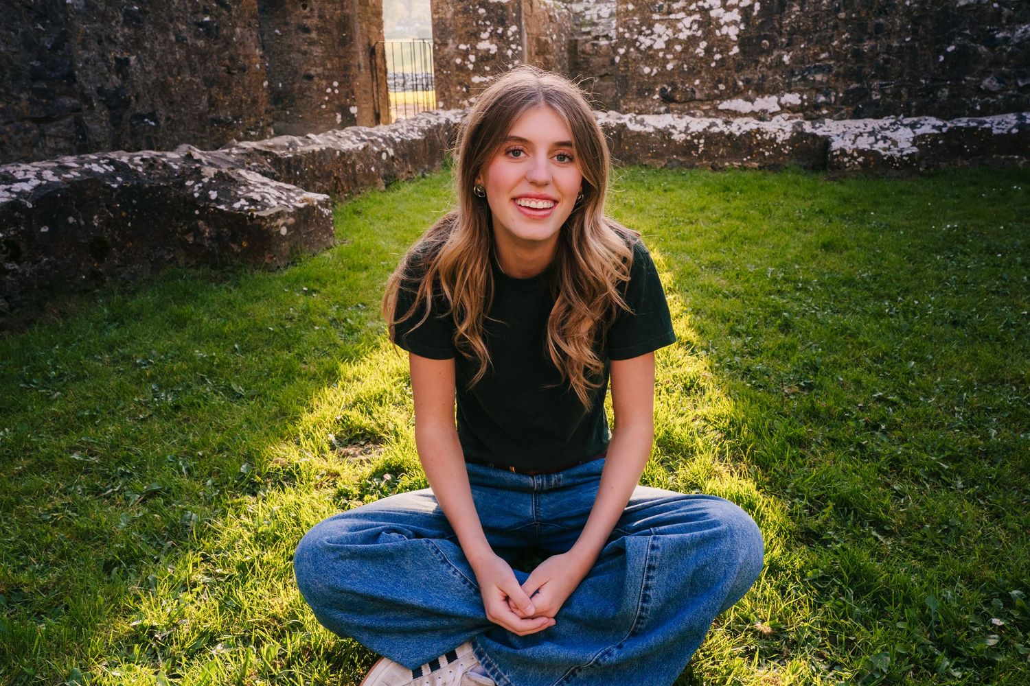 Person in black shirt and jeans sits cross-legged on sunny lawn near stone wall. Photographer Ireland dublin
