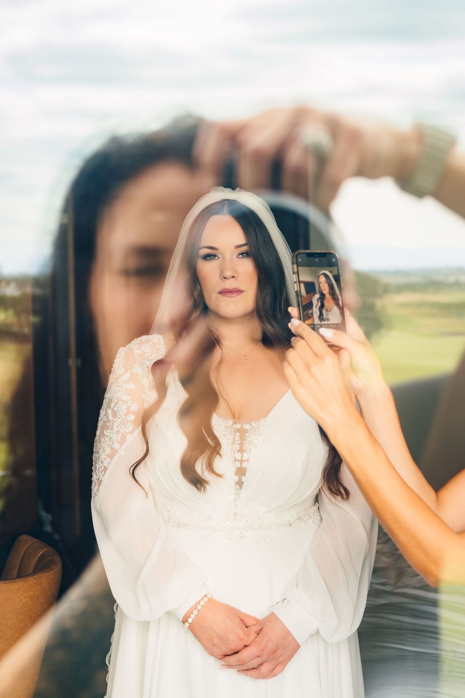 A bride in a flowing white dress with bell sleeves poses while being photographed through reflective glass. Portmarnock Hotel Wedding PHOTOG