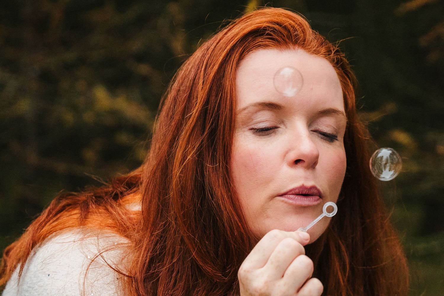 Woman with red hair blows bubbles with eyes closed against dark forest background.