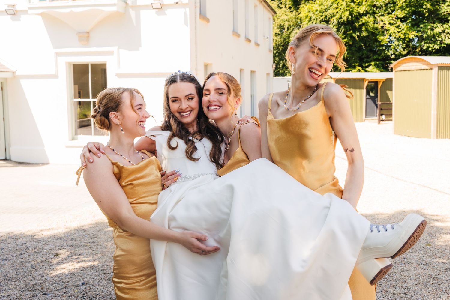 Poulaphouca Wedding photographer wicklow  Bridesmaids in gold dresses lift and carry the bride while laughing outdoors.