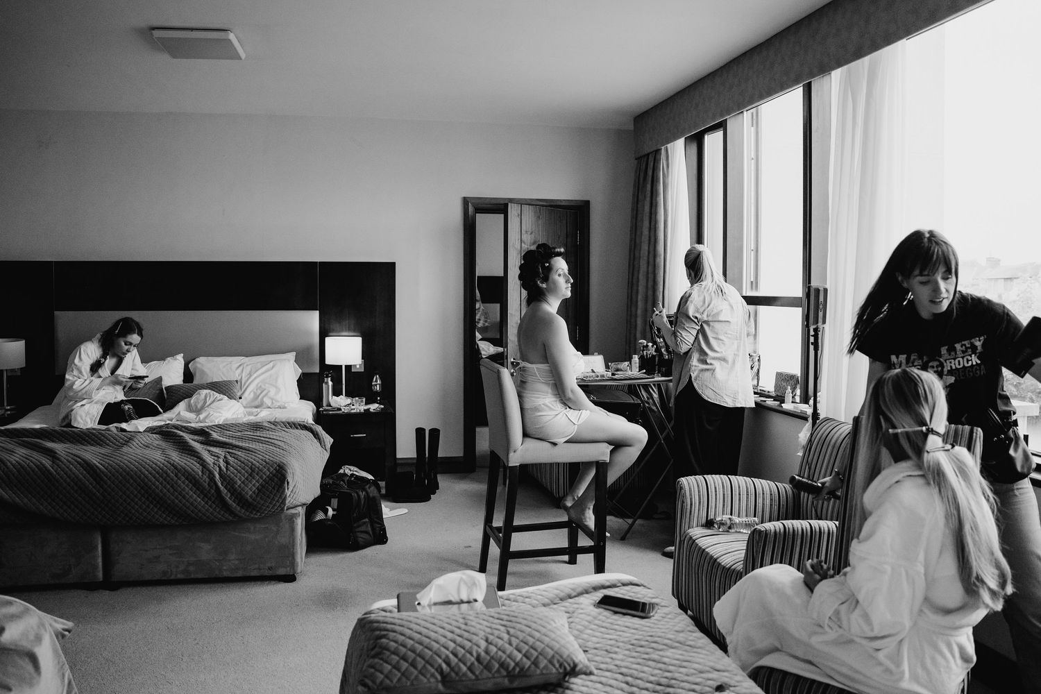 A black and white photo showing people getting ready in a hotel room with natural light streaming through the window. Wedding Photography Tr