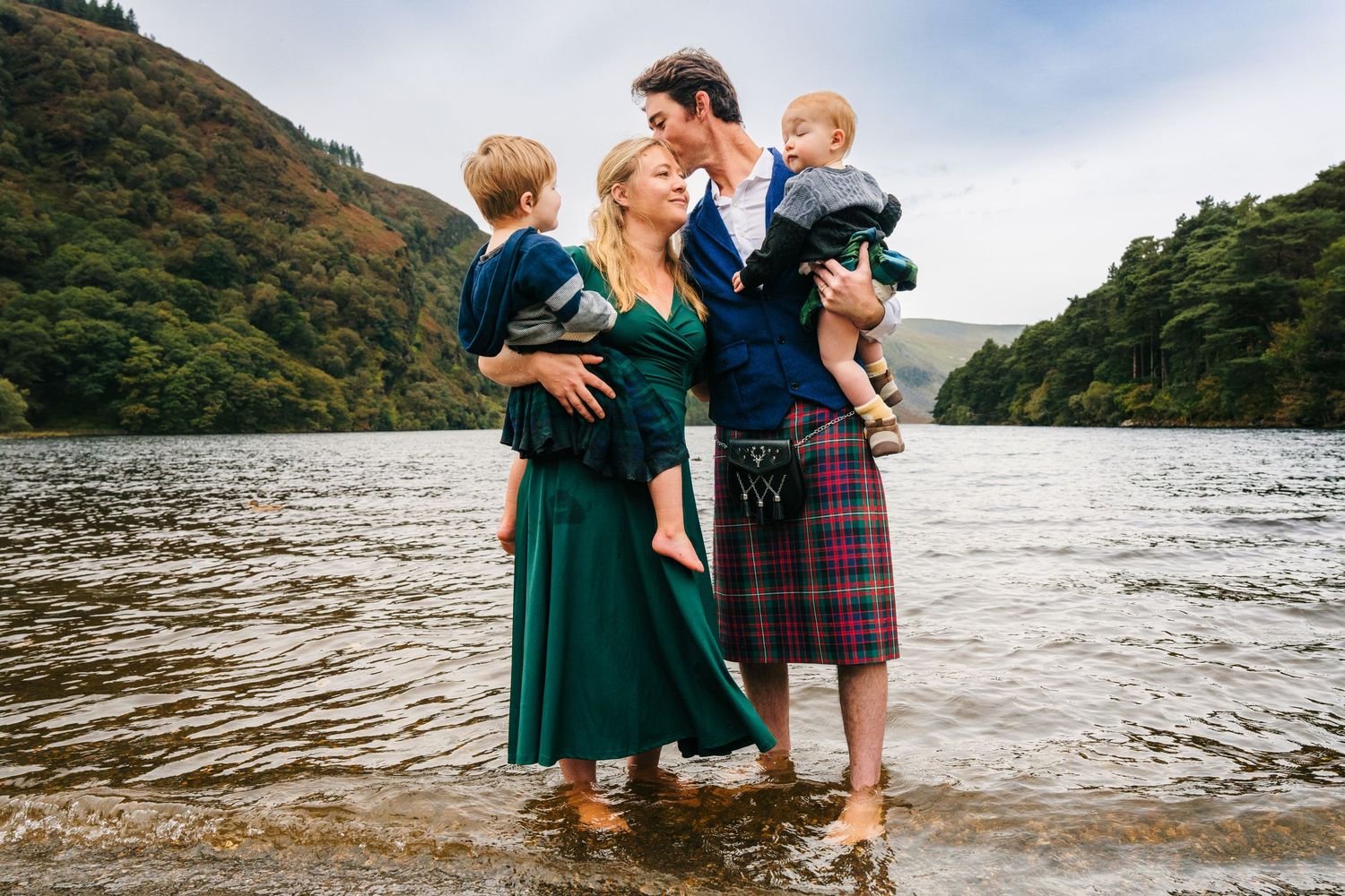 A family wearing traditional Scottish attire stands in a lake with mountains in the background. Photographer dublin ireland