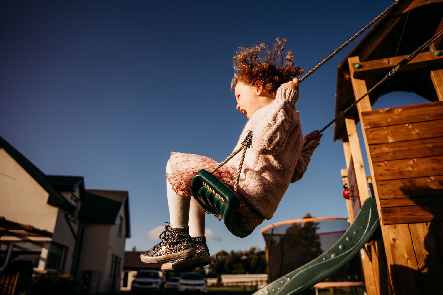 Children playing joyfully on an outdoor wooden swing set at sunset with trees and houses in the background. family PHOTOGRAPHER Dublin
