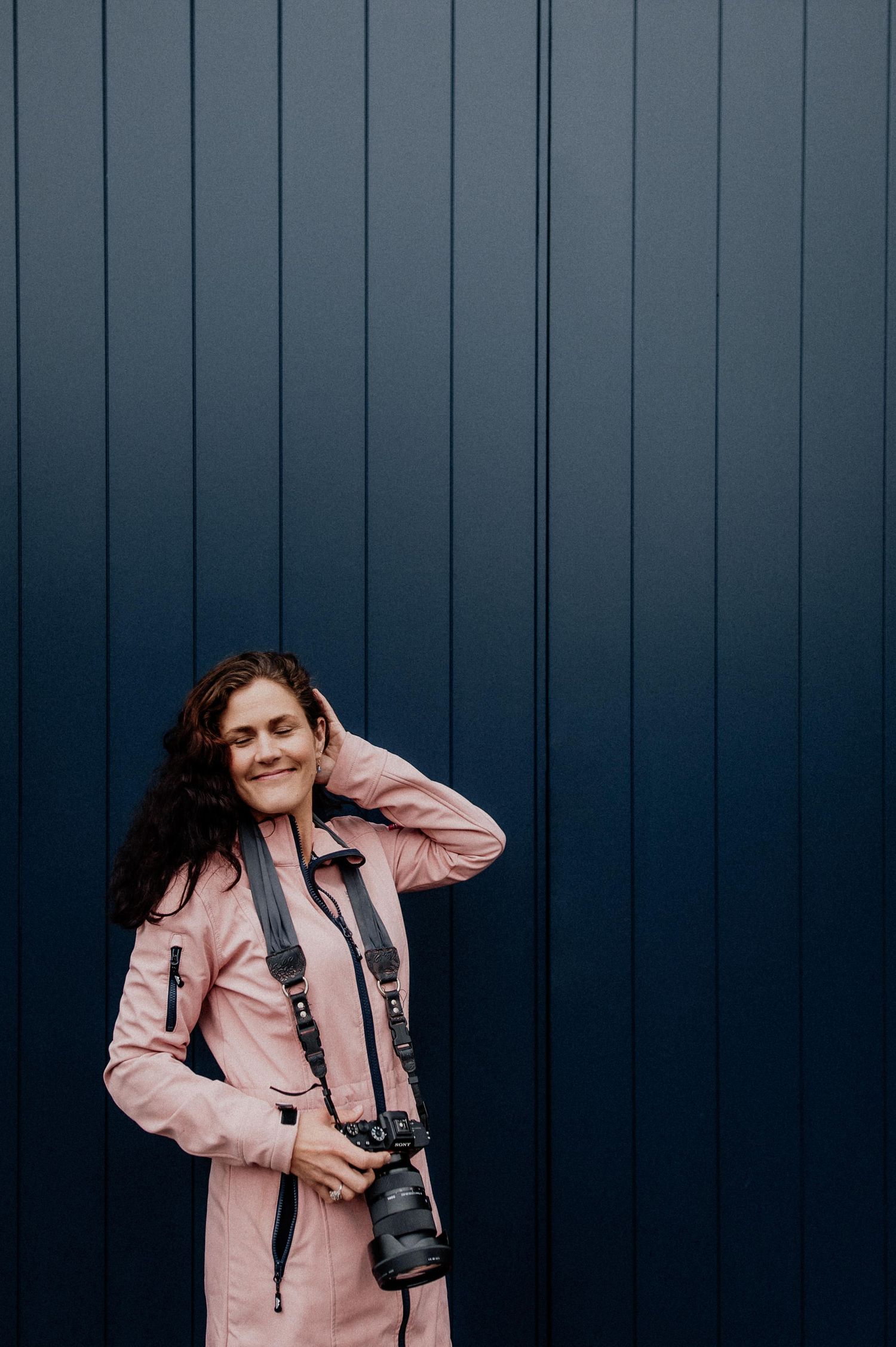 Person in pink coat poses against dark blue wall while holding camera equipment. wedding photographer dublin