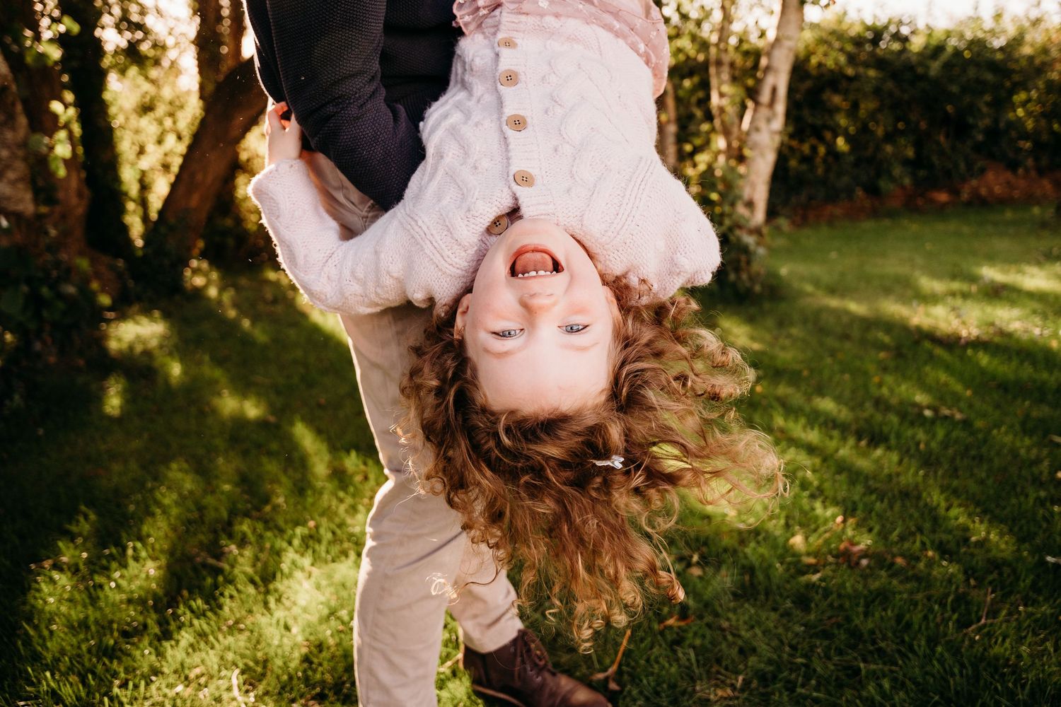 A child hangs upside down from a tree branch in a sunlit backyard, hair flowing freely while laughing and playing. family PHOTOGRAPHER Dubli