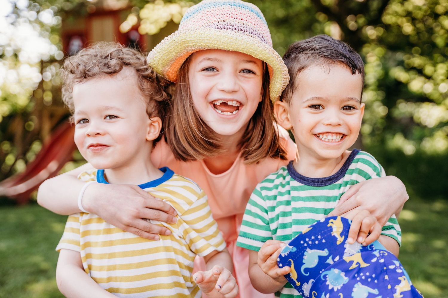 Three young siblings in striped shirts laughing and hugging together outdoors on a sunny day. family PHOTOGRAPHER Dublin