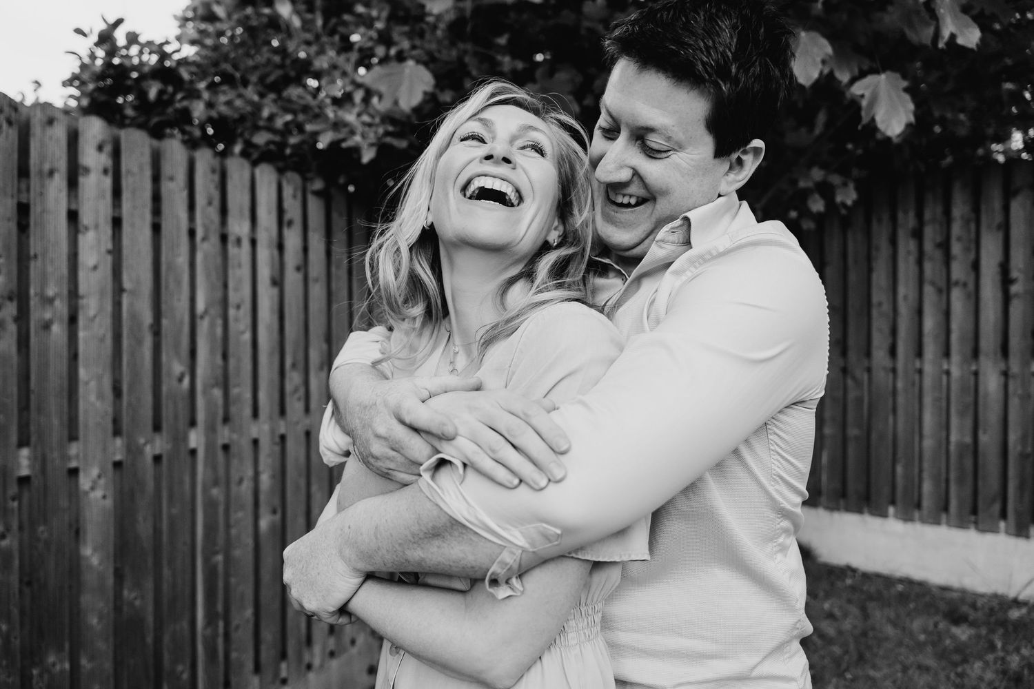 A joyful black and white shot of a couple embracing and laughing together in front of a wooden fence. PHOTOGRAPHER Dublin ireland