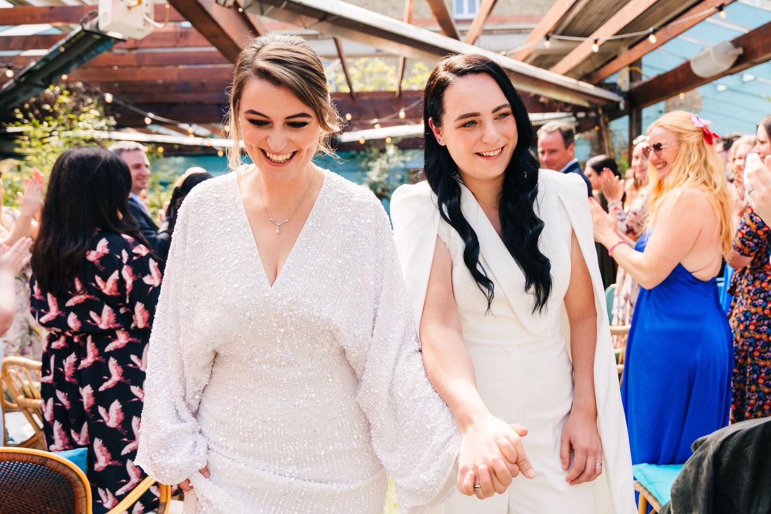 Two people wearing white dresses hold hands while walking together at an outdoor celebration. Wedding PHOTOGRAPHER Dublin
