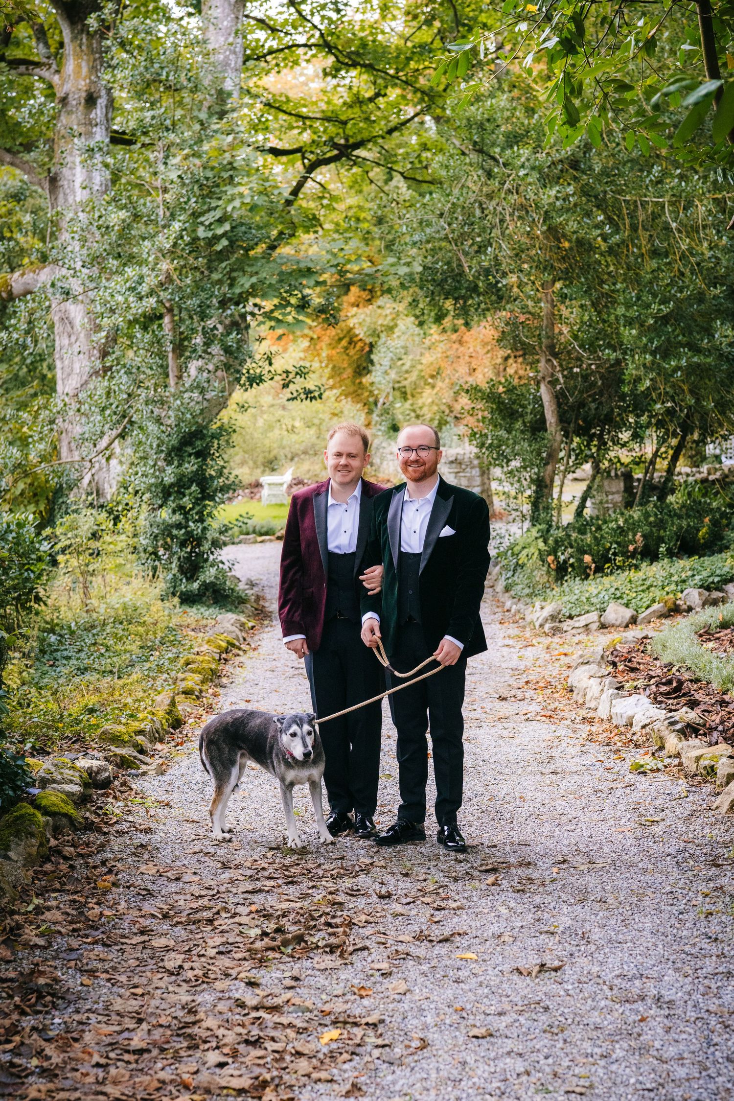 Two people in formal attire walk a dog along a tree-lined autumn path covered in fallen leaves. LGBTQ Ashley Park Wedding Photographer