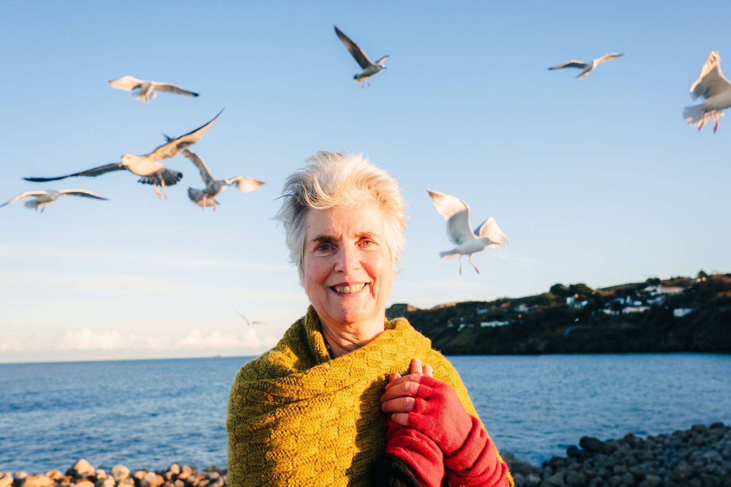 Person in yellow and red winter clothing smiling while seagulls fly around at a coastal location. professional headshots branding photograph