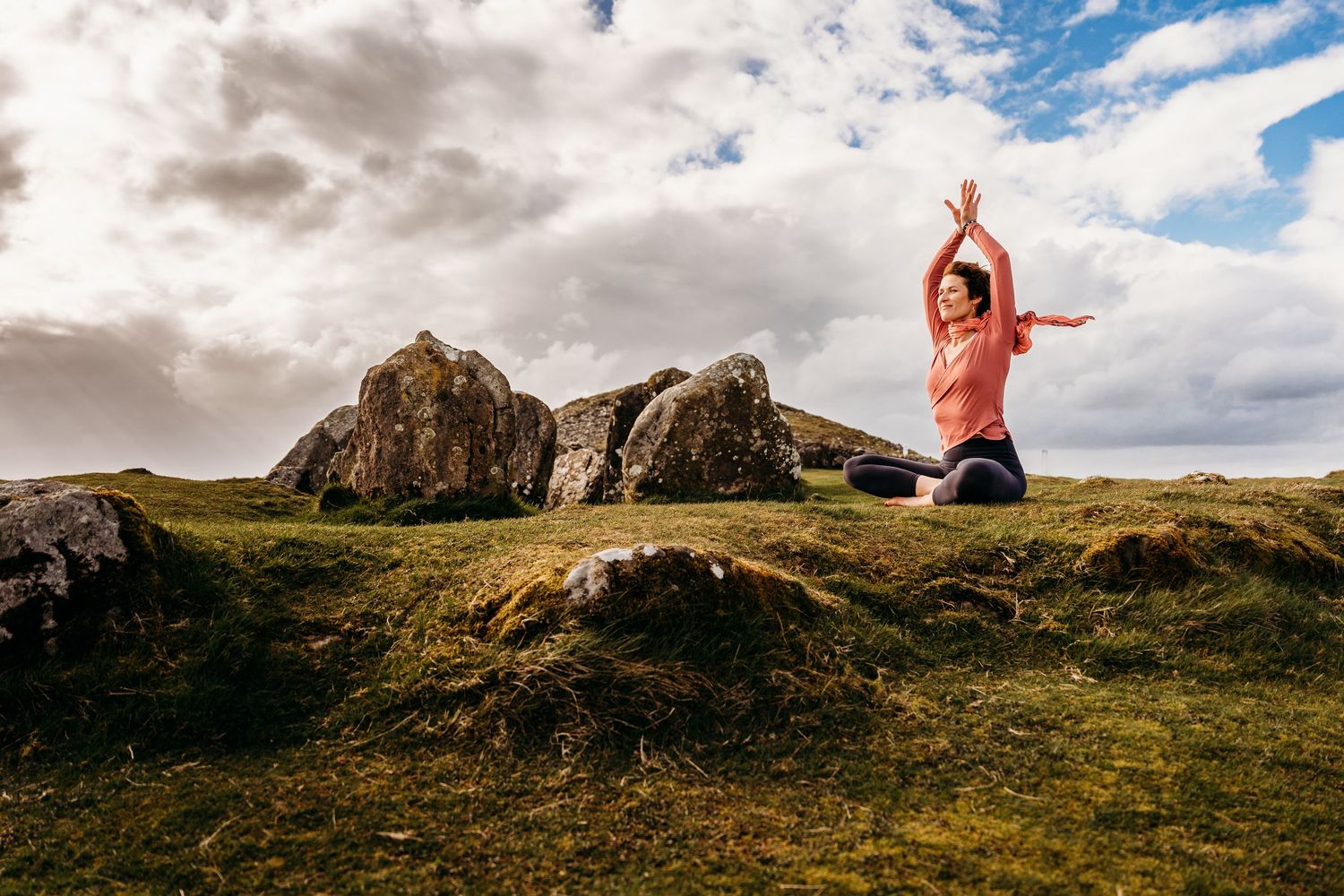 A person in a coral top jumps joyfully against a dramatic mountain landscape with cloudy skies in Scotland. Portrait PHOTOGRAPHER Dublin
