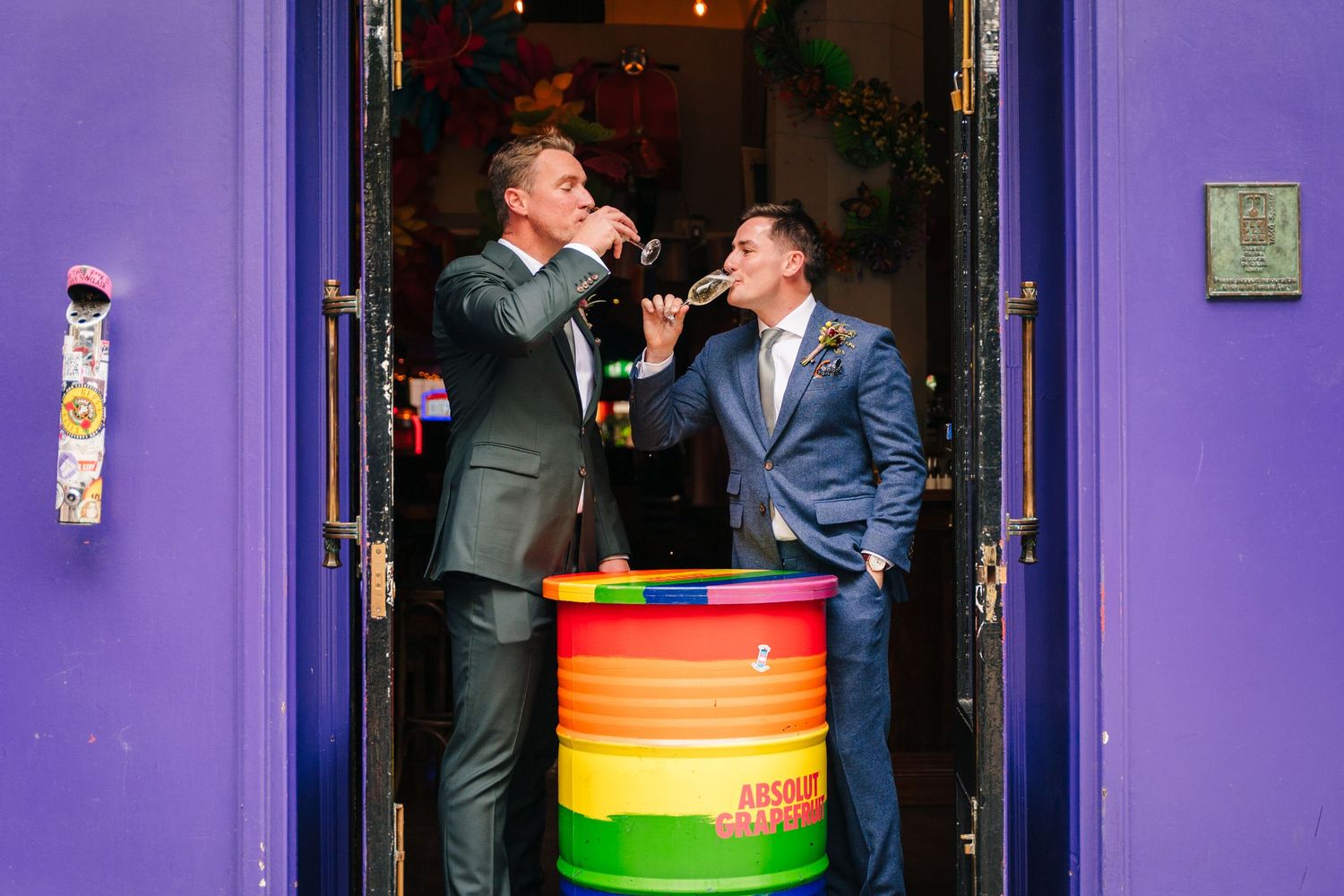 Two people toast with drinks while standing at a colorful rainbow-striped barrel outside a vibrant purple doorway. Same sex wedding PHOTOGRA