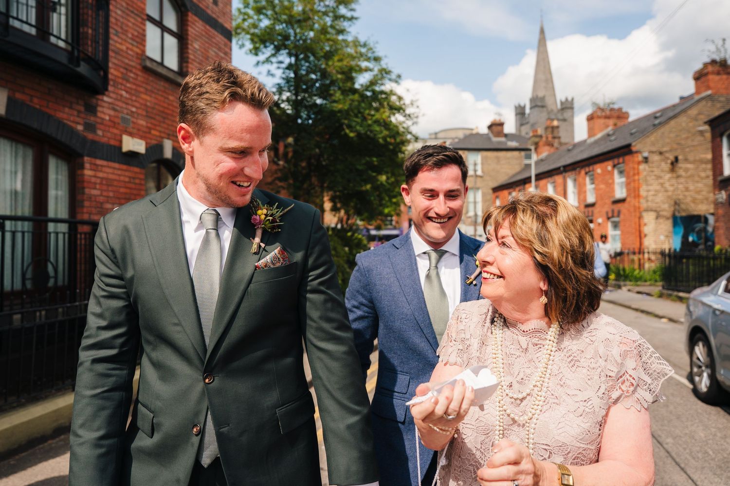 Wedding party walking and laughing together along a brick-lined street with a church spire visible. Same sex wedding PHOTOGRAPHER Dublin