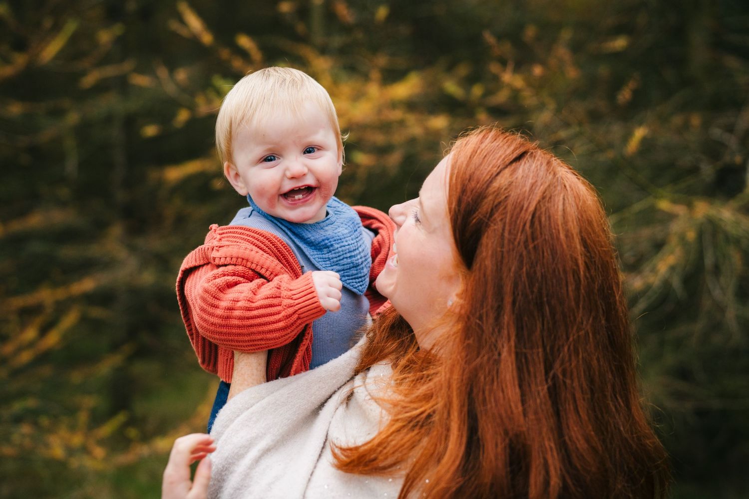 Mother and baby share joyful moment outdoors with autumn foliage in background.