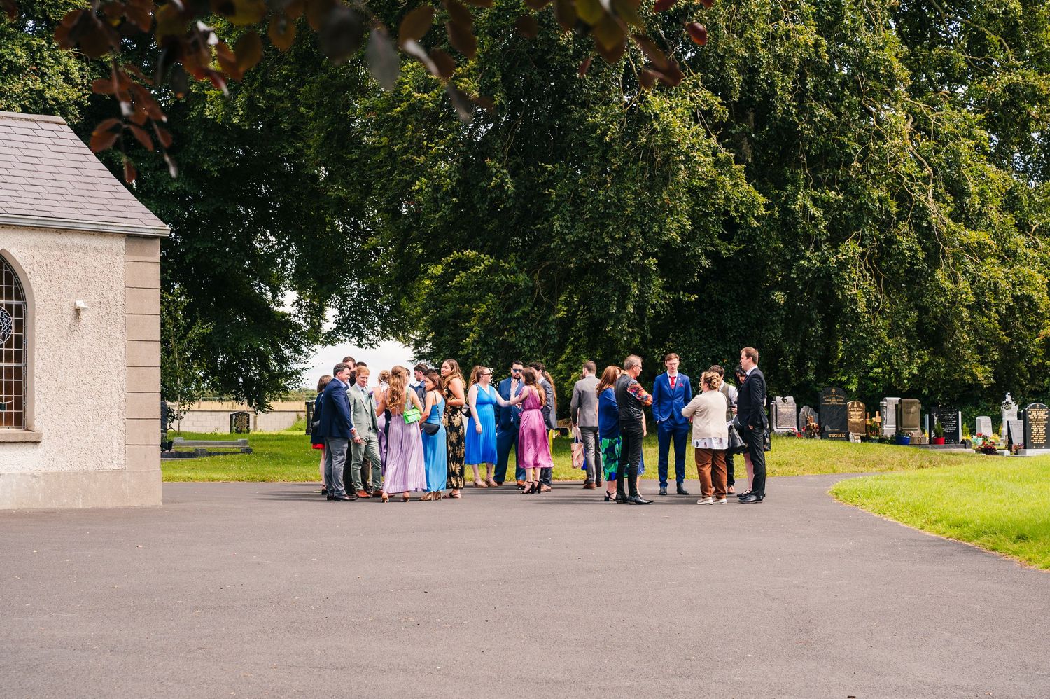 Wedding party gathers outside historic church building on sunny summer day surrounded by trees. Millhouse slane wedding photographer dublin