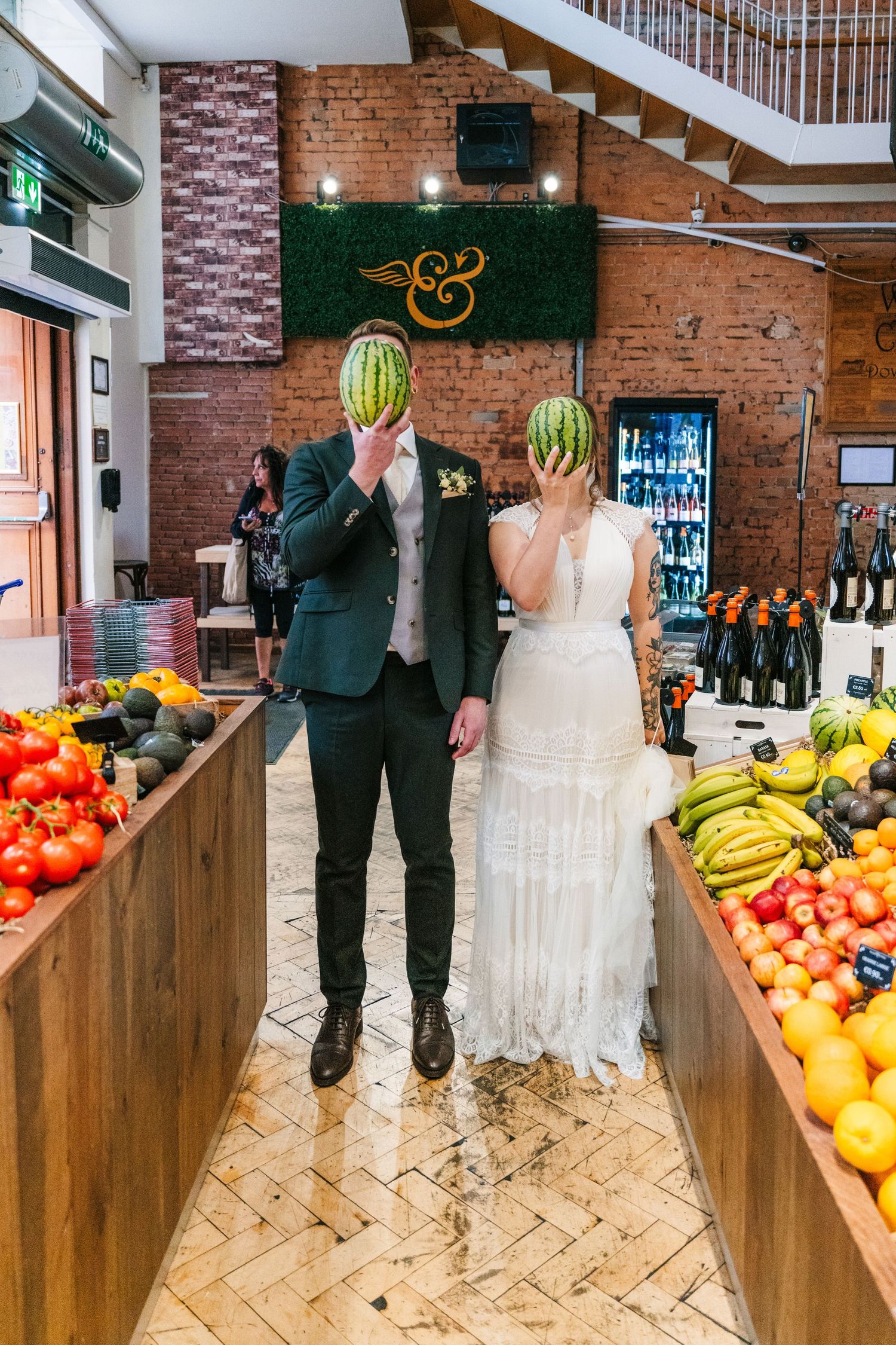 Couple in wedding attire hold watermelons in front of their faces in a produce market. PHOTOGRAPHER Dublin Ireland Wedding