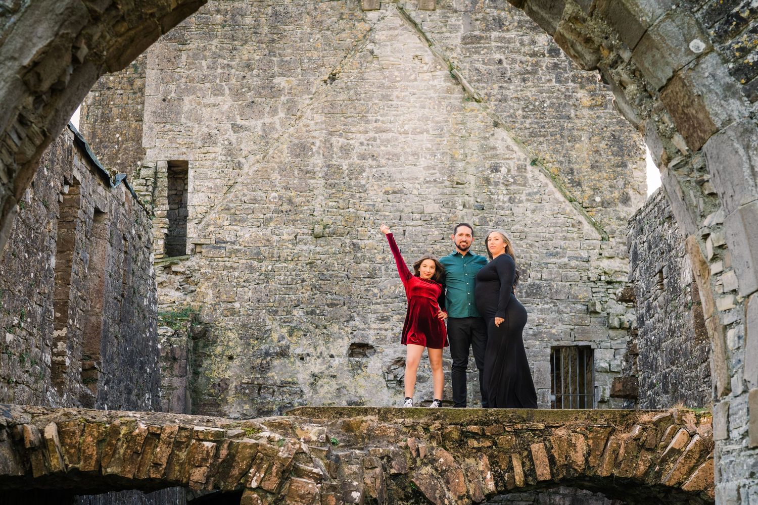 Three people celebrate with raised arms under a stone archway at historic ruins site. Family PHOTOGRAPHER Dublin