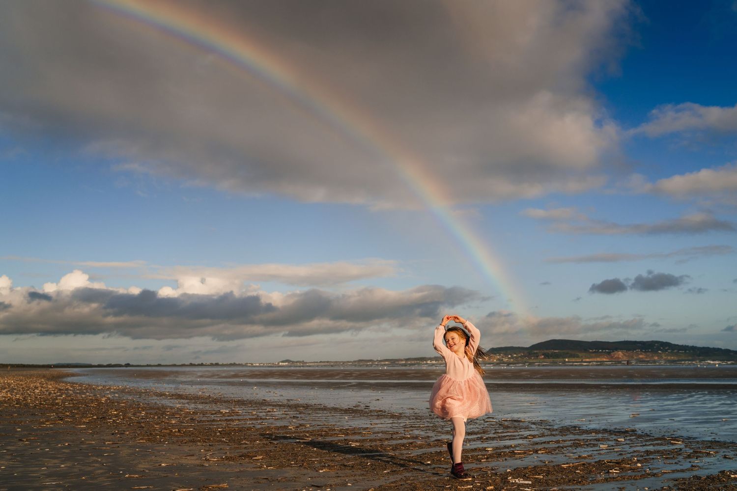 A dramatic rainbow arches over a coastal landscape with rolling hills and scattered clouds in the sky. Family photographer Dublin