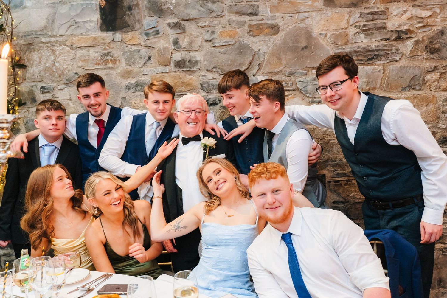 Large wedding party group poses together at reception table against rustic stone wall. Ballymagarvey Wedding photographer