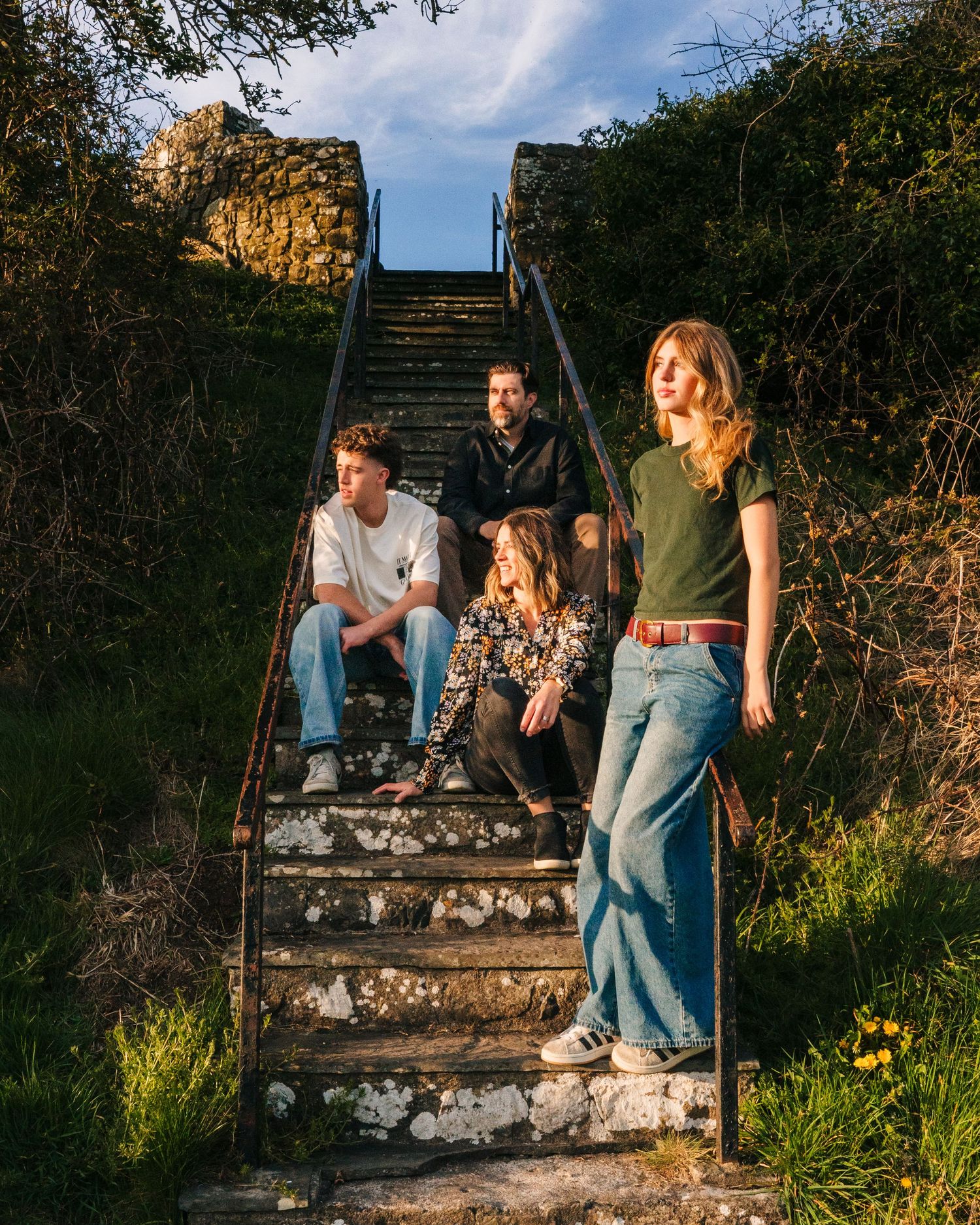 A group of young friends relax on worn concrete steps surrounded by greenery at golden hour as sunlight hits the scene. family PHOTOGRAPHER