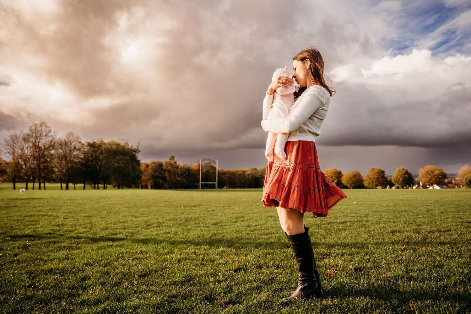 Two figures embrace in a scenic fall field with dramatic clouds and autumn trees in the background. family PHOTOGRAPHER Dublin