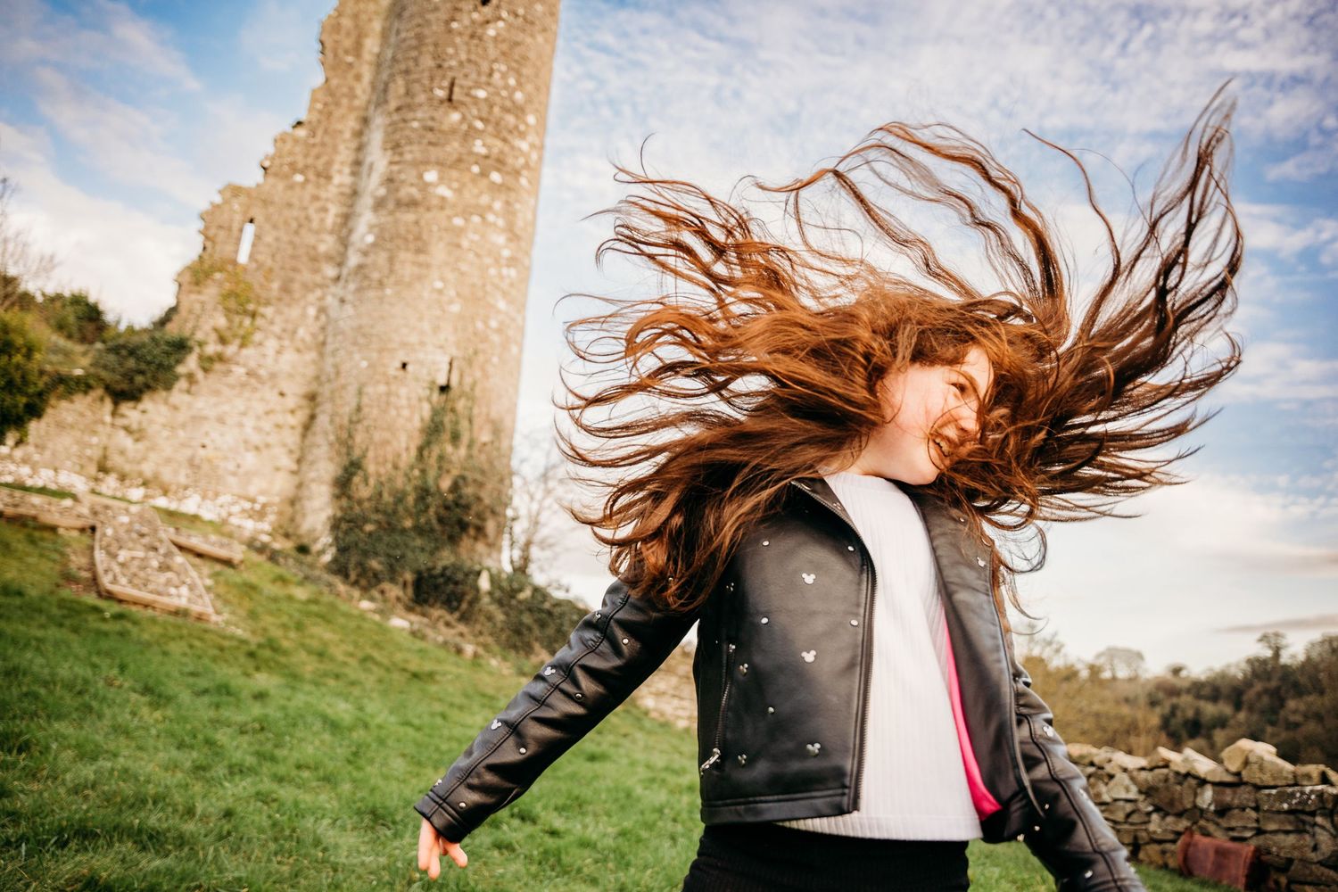 Person with long red hair whipping it dramatically against a stone castle wall backdrop on a cloudy day. family PHOTOGRAPHER Dublin