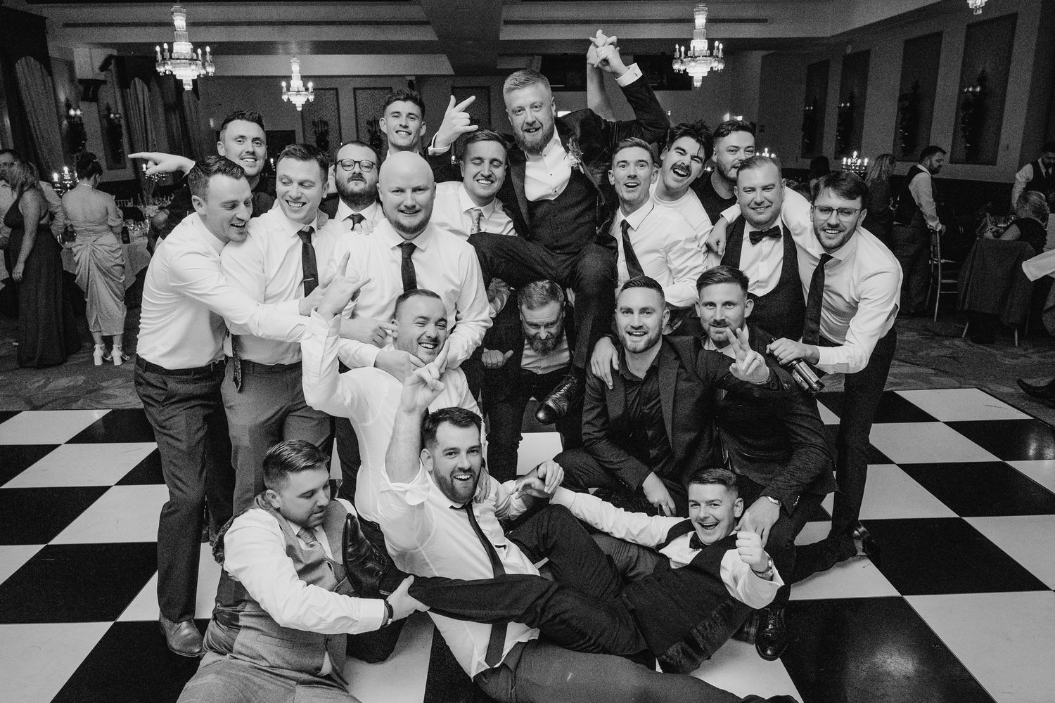 A joyful group of wedding guests gather for a black and white photo on a checkered floor. Wedding PHOTOGRAPHER Dublin
