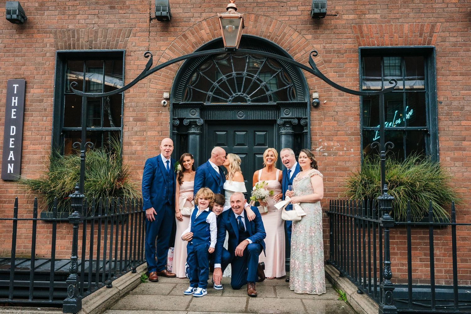 Wedding party poses together along brick wall with black doors and windows at historic venue. The Dean Dublin Wedding Photographer
