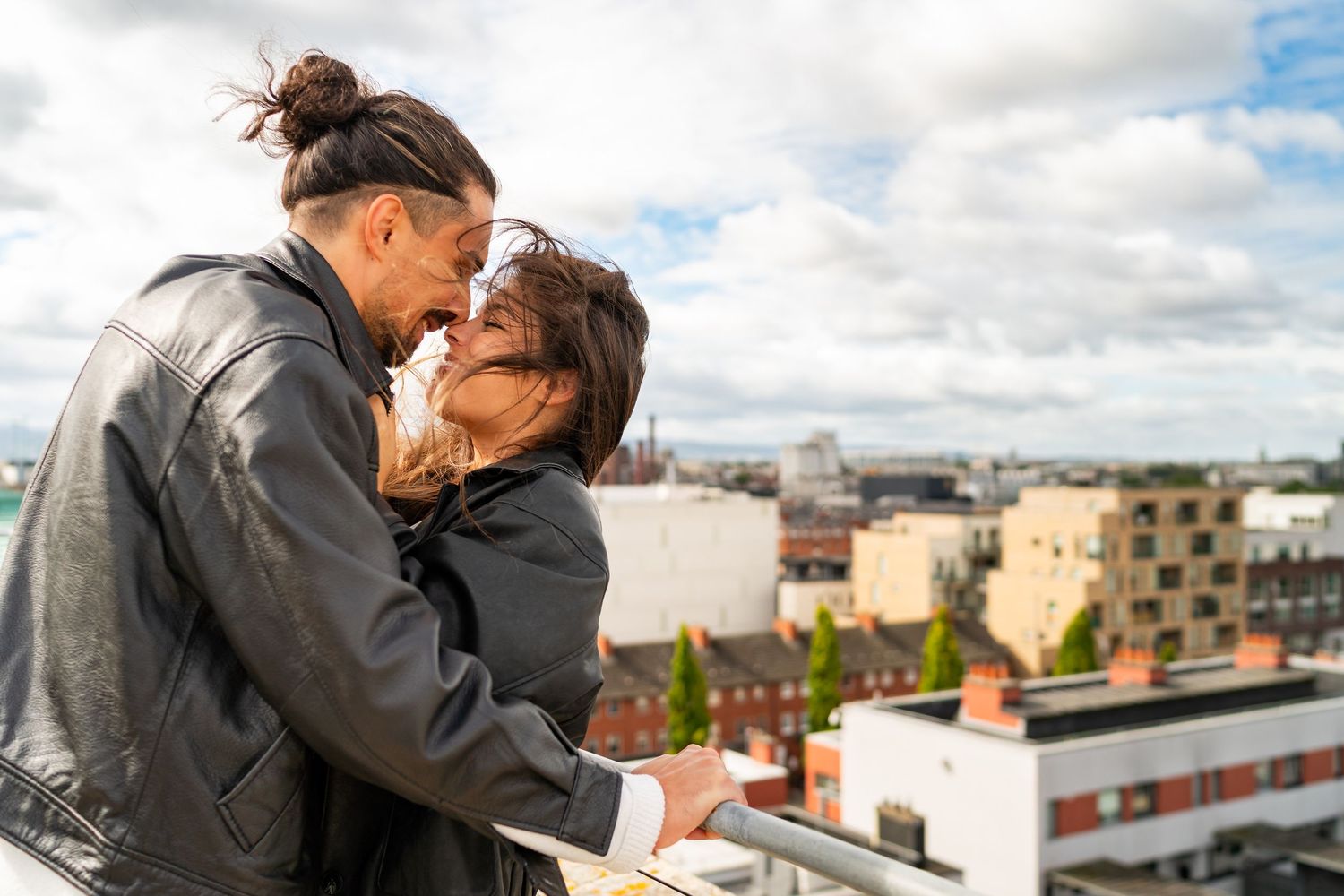 Two people share an intimate moment while standing on a balcony overlooking a cityscape on a cloudy day.