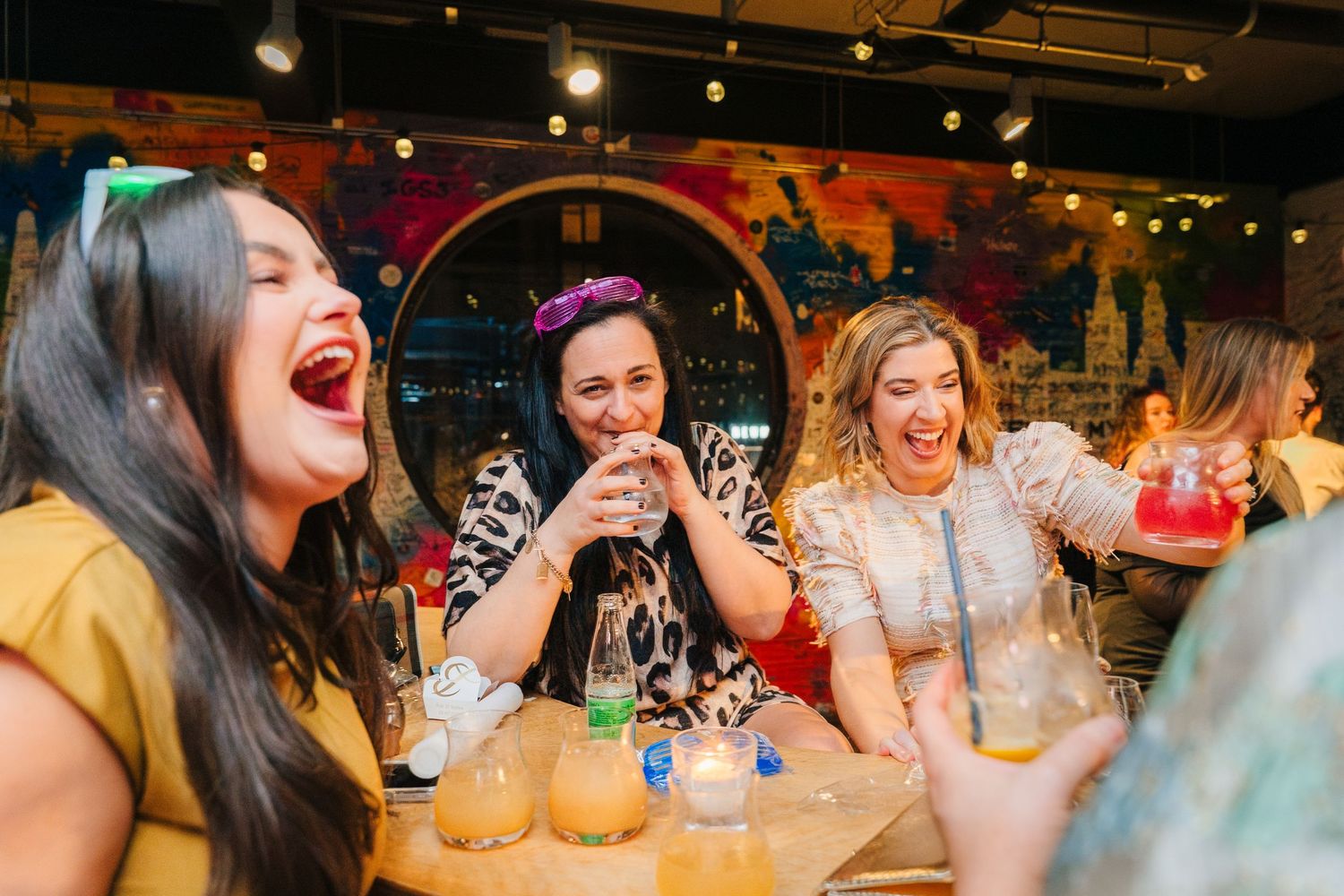 A group of friends sharing laughter and drinks together at a colorful bar or restaurant. Dublin wedding photogrAPher