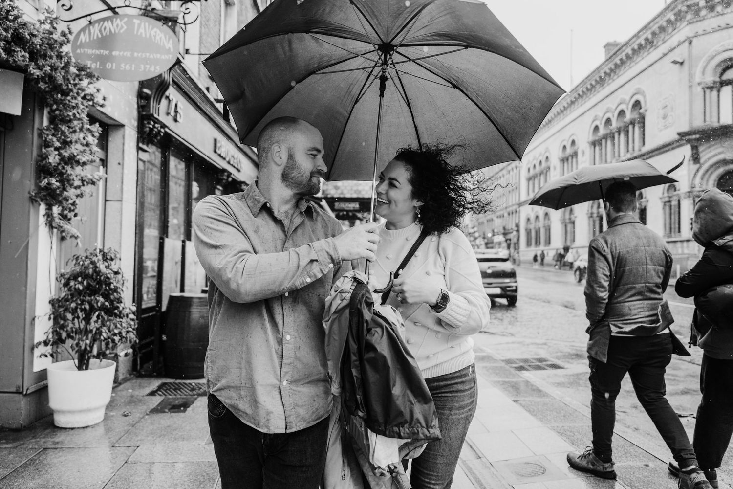 A couple stays dry under a black umbrella on a rainy day in front of urban storefronts. Proposal Dublin Photographer
