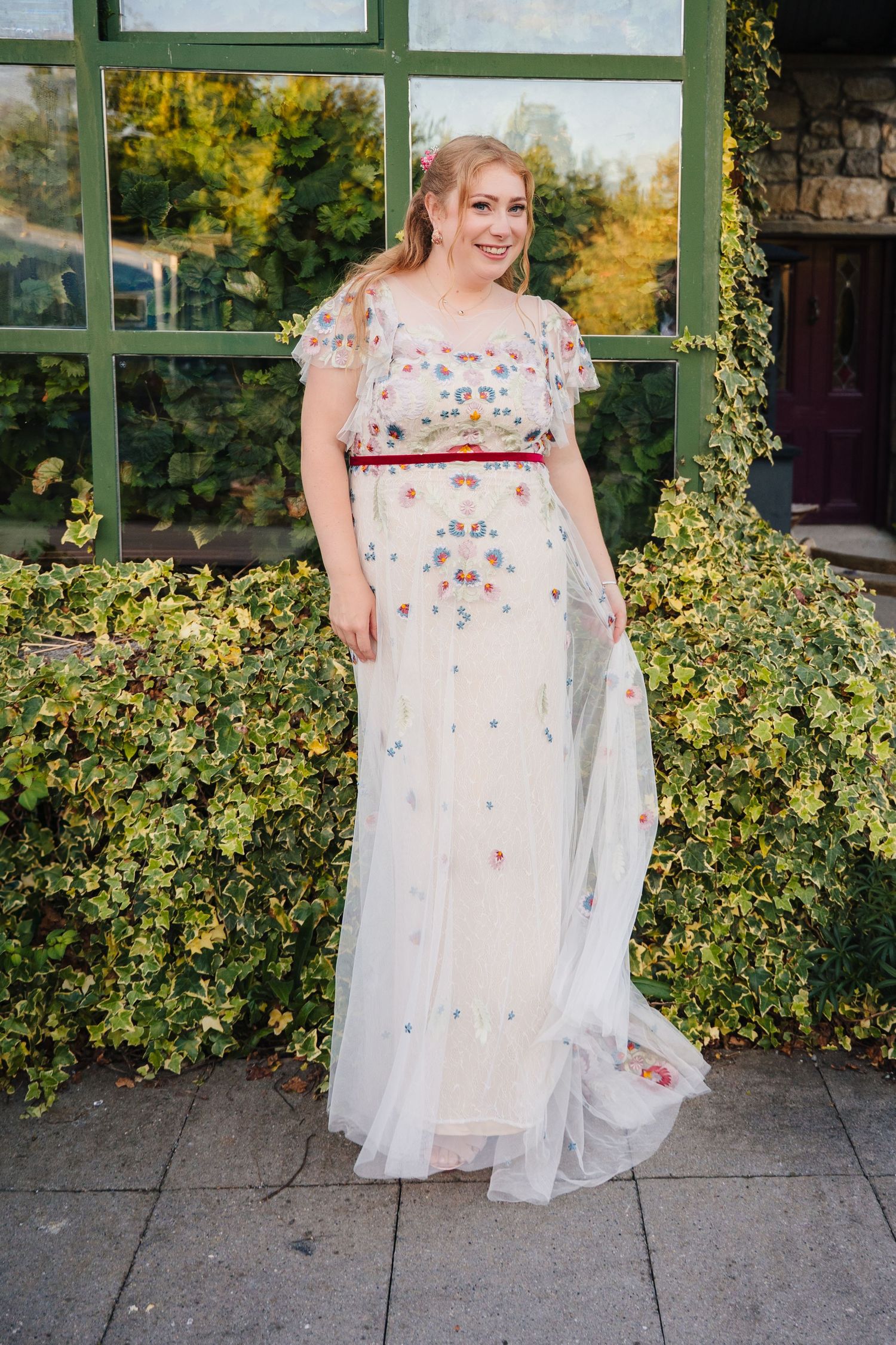 White floral embellished wedding dress with red sash against lush garden backdrop. . Village Barn Tyrrellspass wedding photographer Ireland